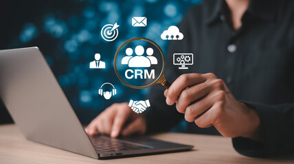  Business concept illustration of a hand holding a magnifying glass over a digital CRM dashboard interface with icons for data analysis, marketing, communication, and customer management.
