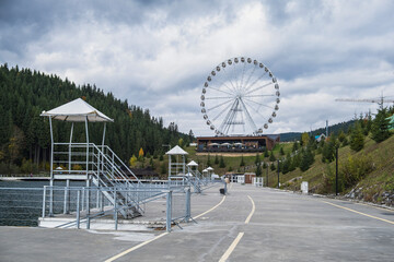 A large white Ferris wheel with enclosed gondolas stands tall against a dramatic sky with blue patches and heavy white clouds, overlooking modern buildings and hills.