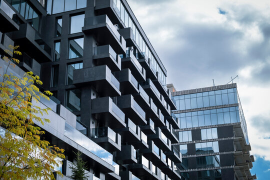 Modern, dark gray facade of a high-rise residential building with projecting balconies and large glass windows, set against a dramatic, cloudy sky, with a touch of autumn foliage.