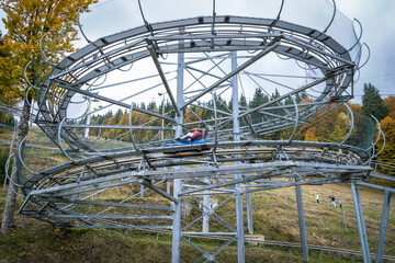 An elevated metal track structure of an alpine coaster (rodelbahn) featuring a looping turn, set on a grassy hill surrounded by a forest with autumn foliage under a cloudy sky.