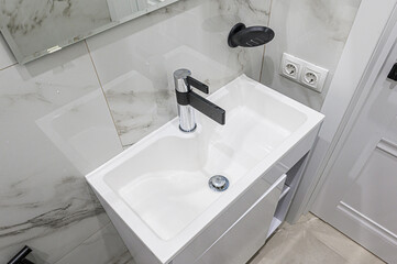 A modern bathroom sink with a black and chrome faucet. The sink is white and set against marble-style tile.  An outlet and soap dish are mounted on the wall