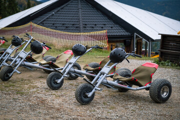 A row of mountain carts with helmets parked at a "Start" gate on a dirt slope near a modern building and a forest, ready for a ride down the hill on a cloudy day.