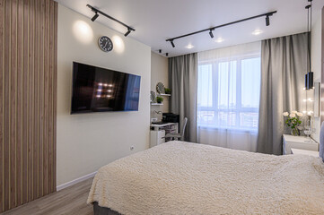 A cozy bedroom features a large bed, TV, workspace, and natural light. The interior is decorated in neutral tones with wood accents and track lighting