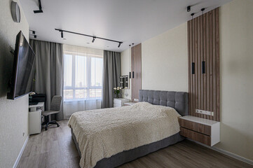 A modern bedroom with a grey bed, cozy blanket, wooden wall accent, and a workspace by the window. Clean, minimalist design