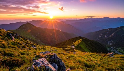 Vibrant sunset illuminates rugged mountain peaks, casting long shadows. Grassy foreground, distant hills fade into the horizon