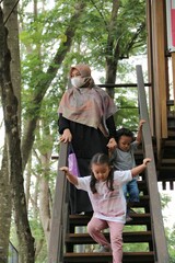 Obraz premium A caring mother wearing a hijab and mask helps two young children descend a wooden staircase in a lush park, conveying family safety, supervision, and outdoor time.