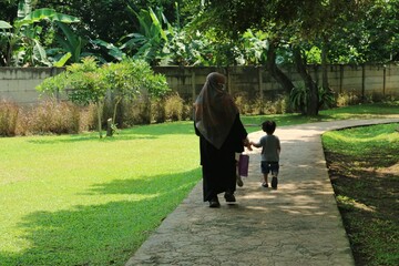 Fototapeta premium Mother Wearing Abaya Walks Hand in Hand With Child Along Park Path Under Green Trees