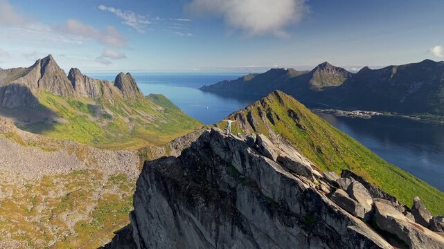 Hiker on rocky peak overlooking calm waters and majestic landscapes Senja island, Norway. Climber celebrating climbing top of Segla mountain by raising his arms 