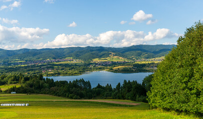 Obraz premium Scenic summer view across czorsztyn lake framed by trees and hills. Small sailboats and green slopes under white clouds emphasize calmness and outdoor leisure.