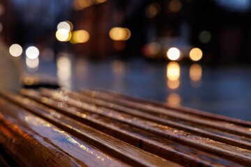 Droplets gather on a polished wooden bench while soft city lights create a warm ambiance on a rainy evening.