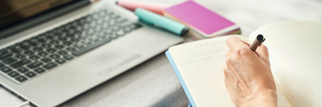 Female hand writing in notebook near laptop with highlighters on table.