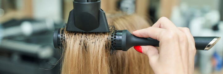 Female hairstyling with round brush and hairdryer: hair care technique for straightening blonde hair in salon setting.