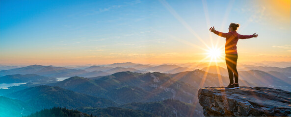 Person celebrating freedom and success at the top of a peak, overlooking rolling hills and a sunburst
