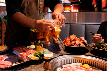 Chefs pour smoky liquid from a playful bull figurine over a feast of meats and vegetables at a...