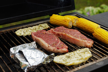Juicy steaks and fresh corn are cooking on a charcoal grill while smoke rises in a sunny outdoor setting.