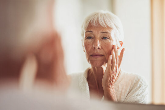 Elderly woman examining her skin in front of mirror indoors