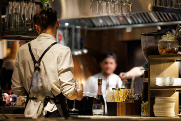 A bartender wearing a white shirt and black apron carefully prepares drinks in a vibrant restaurant setting.