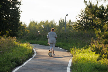 Fototapeta premium A person walks leisurely with a small dog along a winding path in a lush green area during the late afternoon light.