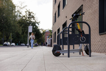 Scooter is secured at a bike rack on a city sidewalk while a woman walks by in casual attire on a sunny afternoon.