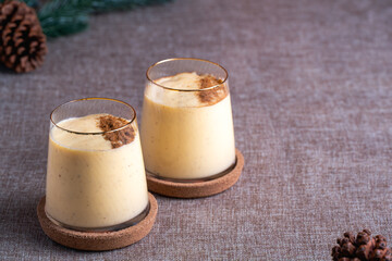 Two servings of creamy eggnog cocktail, dusted with nutmeg, in elegant gold-rimmed glasses on cork coasters over a neutral textile background