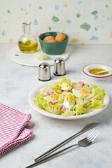 Parisian salad of lettuce, boiled potatoes, ham, and hard-boiled eggs in a light-colored plate against a gray concrete background.