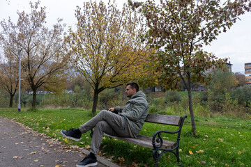 Fototapeta premium A man sits on a park bench surrounded by trees with yellow leaves, wearing casual clothing and enjoying a quiet autumn day in nature, reflecting calmness and outdoor relaxation.