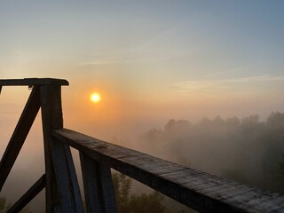 grassy mountain slope with a wooden fence, lit by morning sun. Tall evergreen overlooks hazy valleys. moody winter sunset over the Cotswold countryside. Sunlight on the morning with sea of fog.