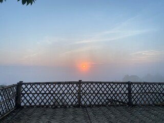 grassy mountain slope with a wooden fence, lit by morning sun. Tall evergreen overlooks hazy valleys. moody winter sunset over the Cotswold countryside. Sunlight on the morning with sea of fog.