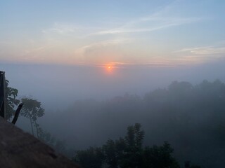 grassy mountain slope with a wooden fence, lit by morning sun. Tall evergreen overlooks hazy valleys. moody winter sunset over the Cotswold countryside. Sunlight on the morning with sea of fog.