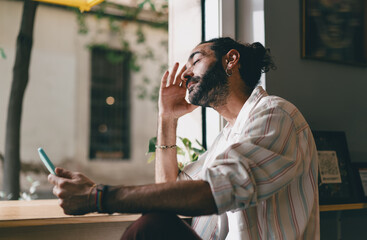 Young Middle Eastern man sitting by window holding smartphone, eyes closed, expressing digital fatigue or screen burnout, showcasing emotional impact of constant connectivity.