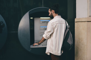 Man with backpack stands at ATM, entering information, representing financial self-reliance, outdoor banking and digital transactions in modern independent lifestyle. © BullRun