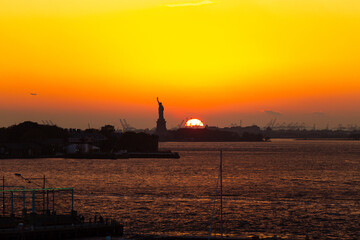Sunset over New York Harbor with the Statue of Liberty silhouetted against the vivid orange and yellow sky with an airplane flying in the distance