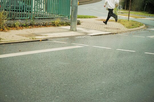 Mann mit schwarzer Hose und wei&szlig;em Anorak l&auml;uft mit gro&szlig;en Schritten &uuml;ber Ampelkreuzung von Stra&szlig;e auf Gehweg in Stadt bei Regen am Nachmittag im Herbst