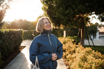Smiling senior woman 55-60 year old wearing eye glasses walking in city street outdoors over sun light. Happiness.