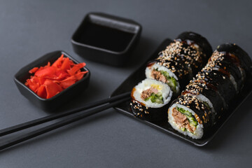 Sushi roll with salmon,  avocado and tuna wrapped in nori seaweed. Sushi set served on the black plate next to red ginger with black sticks on the black background. Flat lay