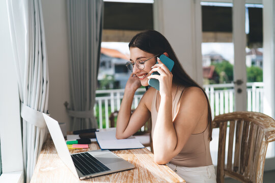Freelancer woman multitasking with phone and laptop smiles during remote conversation, showcasing efficient digital workflow and communication in home-based work life.