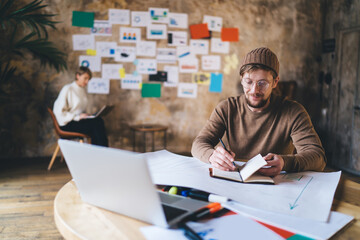 Smiling freelancer takes notes at desk with laptop and diagrams, highlighting joy in digital creation, startup success, and autonomy in modern remote tech-driven careers.