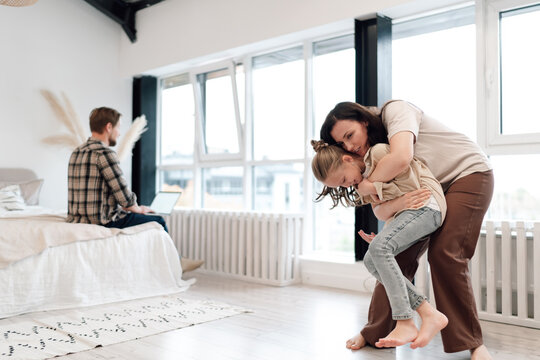 Caucasian man working on laptop while sitting on bed in background, as mother lifts daughter playfully in foreground—contrasting work from-home tech with family bonding.