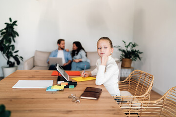 Daughter looks away from her work with distant expression while parents focus on each other,...