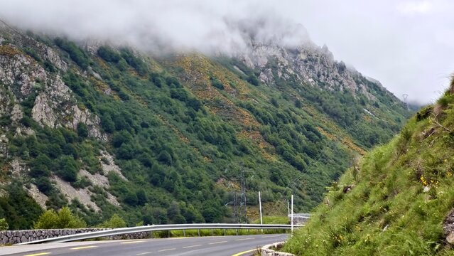 Curving mountain road in a lush, green landscape with steep slopes covered in dense vegetation and rocky outcrops The road is bordered by a metal guardrail. Thick clouds partially obscure the mountain