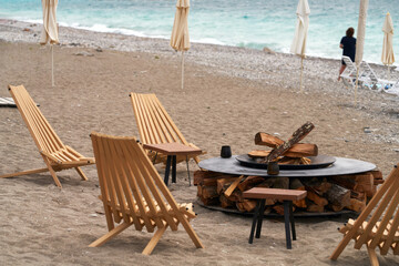 A portable fire pit with wood on the seashore. Wooden lounge chairs surround the circular metal structure. An evening beach retreat by the fire.
