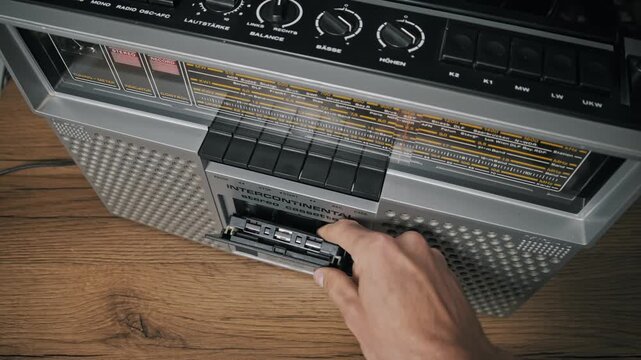 High-angle perspective of a hand pressing the eject button on a vintage silver boombox, opening the deck door to remove an old audio cassette with a yellowed label.