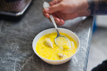 Close-up of Tattooed Hand Scooping Creamy Yellow Chicken Soup with Spoon from White Bowl on Gray Marble Kitchen Countertop