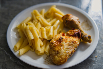 Plate of Golden Roasted Chicken Drumstick Served with Buttered Penne Pasta on White Dish atop Gray Marble Table Surface in Home Setting