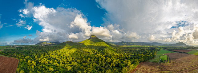 Mauritius volcanic peak from drone over lush forest valley, sugarcane fields, dramatic clouds and sunlit slopes © ronedya