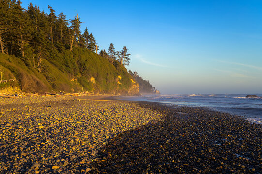 Ruby Beach shows a pebble shore with steep, forested cliffs under clear evening light in Washington State.
