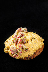 Gourmet pastry topped with piped yellow cream and decorative white chocolate bark with dried fruits and nuts. Elegant dessert presentation on a neutral background.