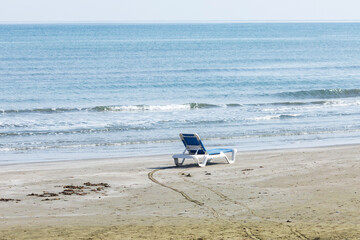 A single, empty white and blue beach lounger rests on a wide stretch of sandy shore. The empty lounger symbolizes a peaceful invitation to relaxation and off-season solitude.