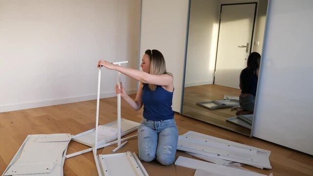 Woman assembling white flat-pack furniture on wooden floor in minimal room. DIY home setup, moving in, hands-on interior work. Mirror reflection adds depth to cozy indoor scene.