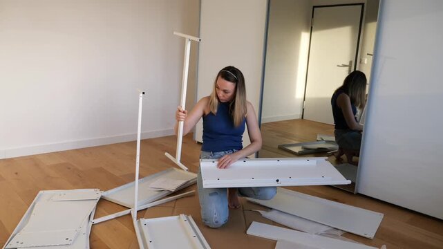 Person assembling white flat-pack furniture on wooden floor in minimal room. DIY home setup, moving in, hands-on interior work. Mirror reflection adds depth to cozy indoor scene.
